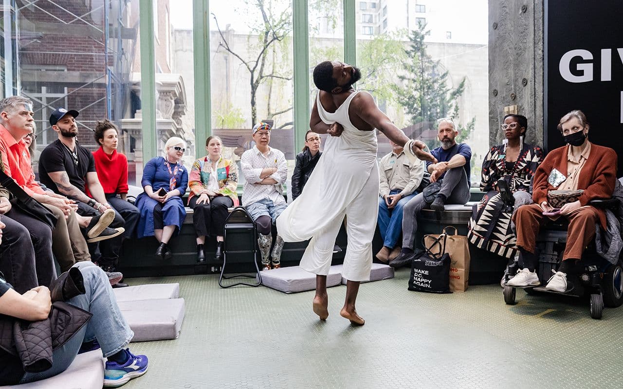Jerron twirls in mid turn dressed in a long flowy white tunic in front of audience in a light filled atrium. Photo by Liz Ligon